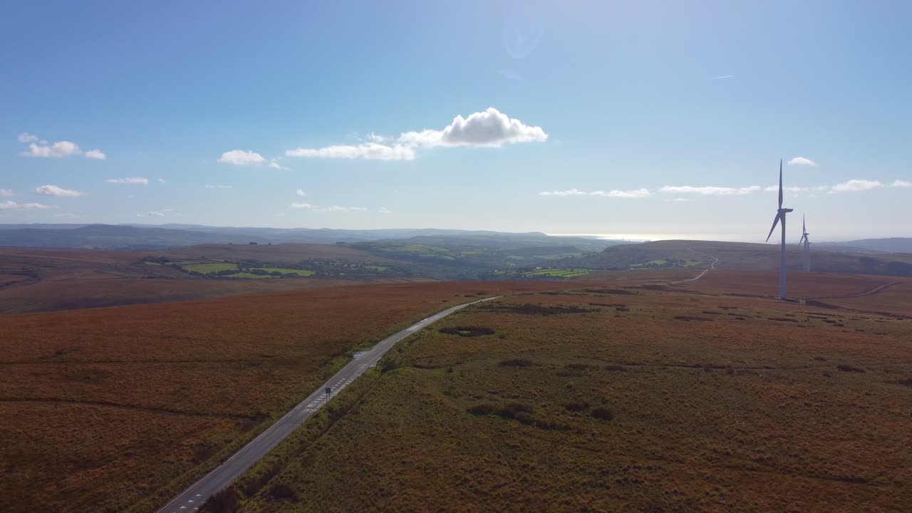 Descending Aerial Drone on Moorland with Wind Turbines and Long Straight Road with Fields and Blue Sky Background 4K