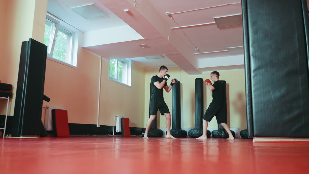 Two boxers sparring in martial arts gym practicing fighting stance with gloves on red mat floor surrounded by punching bags under daylight from windows during training session focused on combat sport
