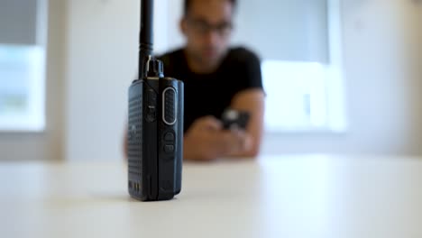 una radio bidireccional colocada en una mesa blanca con una persona borrosa sosteniendo un dispositivo en el fondo, destacando el concepto de comunicación y tecnología en un entorno moderno
