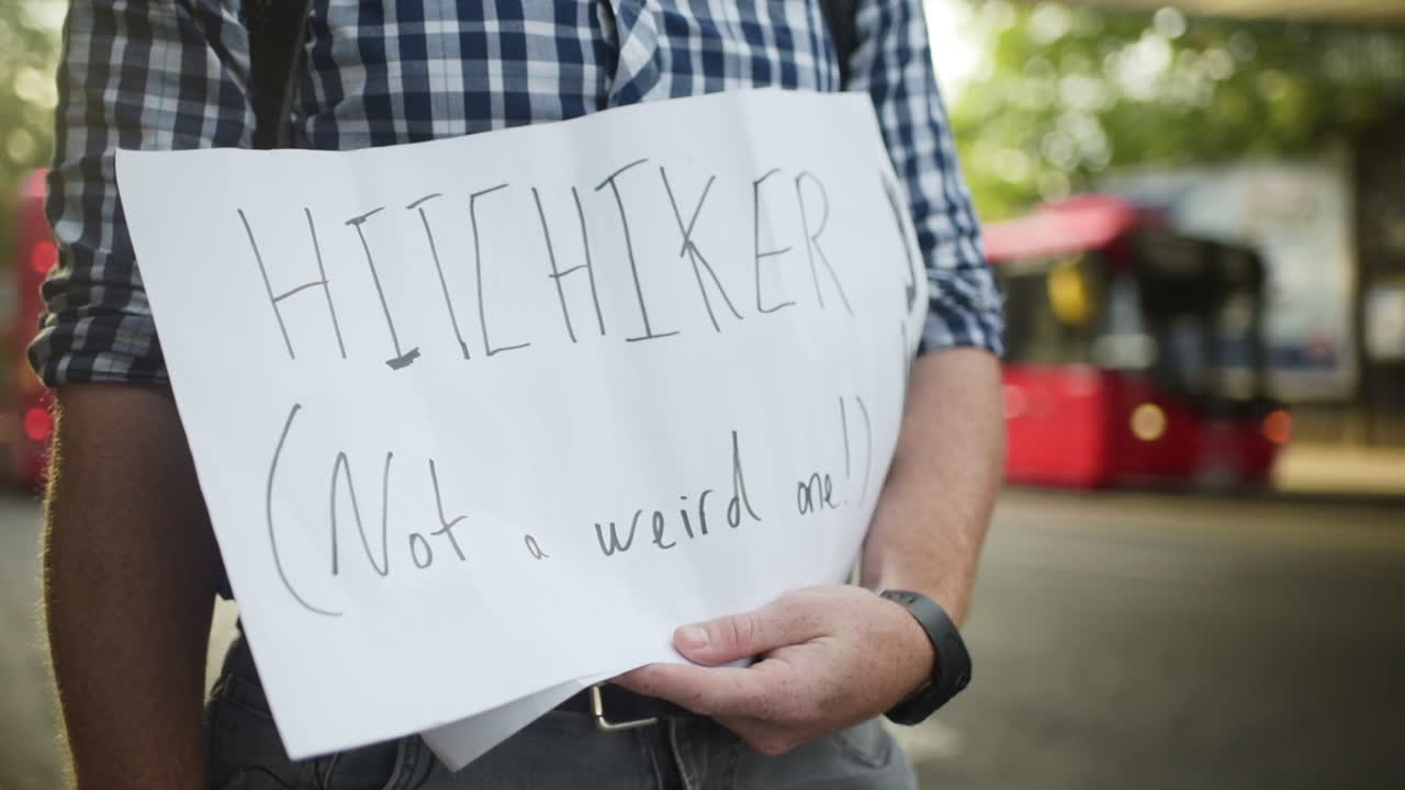A hitchhiker holds a sign up asking for rides on the side of the street with cars and busses passing by in the blurred background