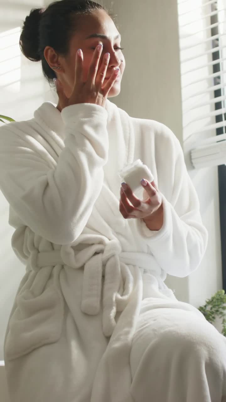 Vertical video: Light urging lady in robe dipping into jar rubbing cream on face at window for skin