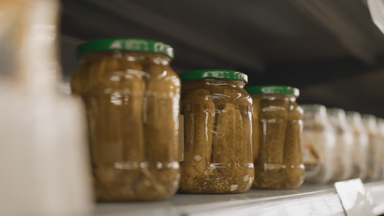 Close up of tattooed hand grasping glass jar of pickles from supermarket shelf in bright grocery aisle highlighting green lid polished nails and retail selection with blurred rails and setting