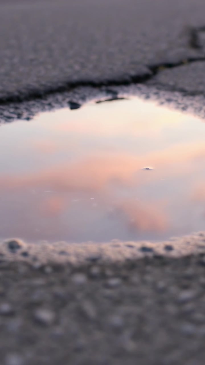 Vertical video: Focusing low camera on hole reflecting pink sky cracked road pebble drifting