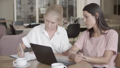 focused blonde woman writing something on paper with pen while her coworker looks at ther