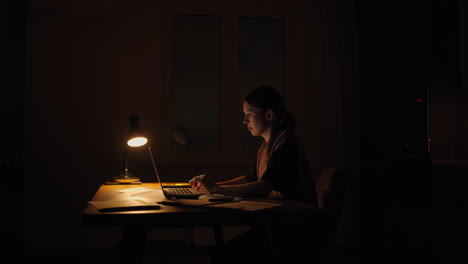 una mujer por la noche bajo una lámpara trabaja desde casa en la oscuridad. hora de la noche para el trabajo plazos son plazos de quema.