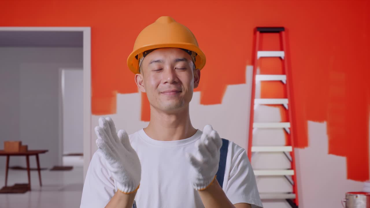 Close Up Of Asian Man Painter Wearing Safety Helmet Smiling And Clapping His Hands While Standing With Painting Wall In Room