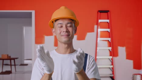 un pintor asiático que usa un casco de seguridad sonriendo y aplaudiendo sus manos mientras está de pie pintando la pared en la habitación