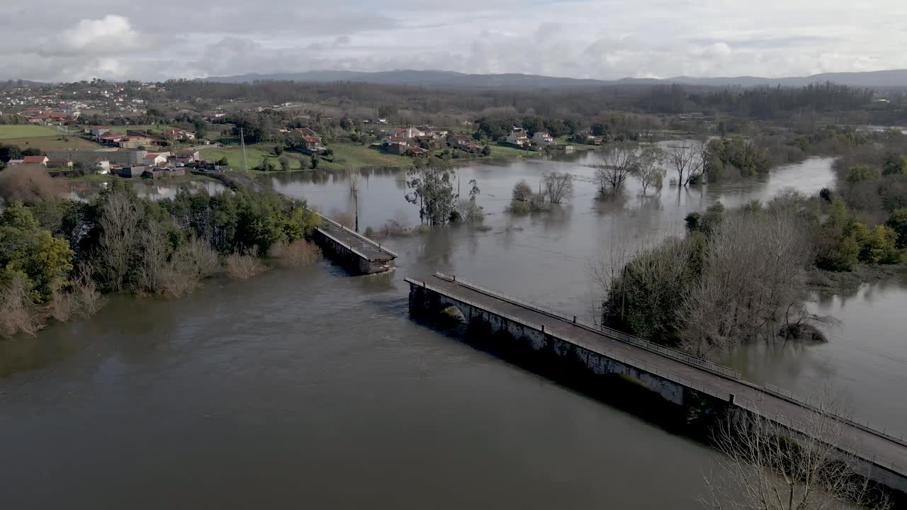 A perspectiva aérea enquadra a travessia quebrada dentro do vale alagado mais amplo, mostrando como a expansão do rio impactou diretamente a estrutura da ponte. Drone girando para a esquerda mostrando a ponte quebrada