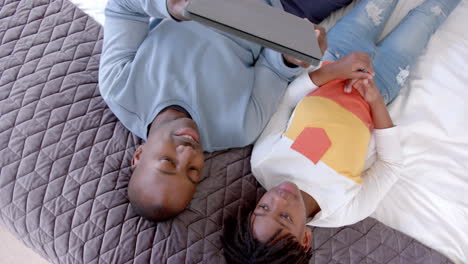 happy african american father and son lying on bed using tablet at home, slow motion