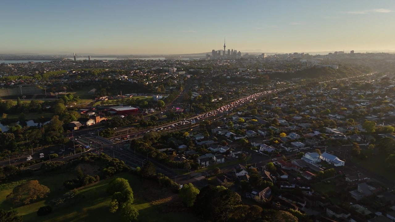 Un'autostrada trafficata si snoda oltre il campo da golf Chamberlain Park verso lo skyline di Auckland al tramonto in Nuova Zelanda. La Sky Tower si erge sopra i sobborghi nebbiosi vicino all'acqua calma di uno stagno. Ripresa aerea con drone in avvicinamento.