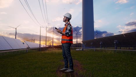full body side view of asian male engineer with safety helmet using smartphone and looking around while standing with solar panel and wind turbines