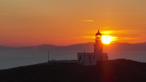 cinematic drone shot of armenistis lighthouse in mykonos during golden hour as the sun sets on the horizon, casting warm light over the aegean sea