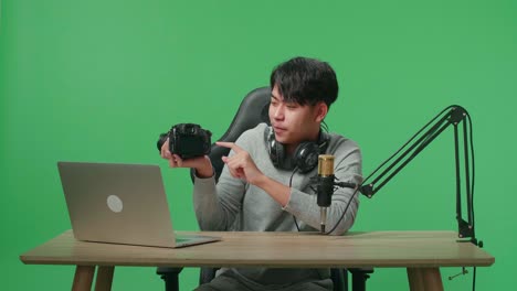 a smiling asian man with headphone and computer reviewing camera while sitting in front of green screen background
