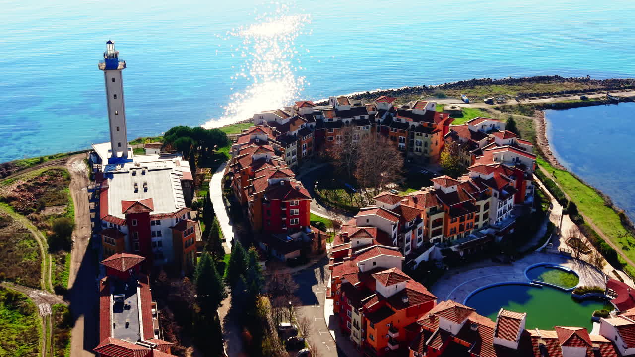 Ripresa aerea dall'alto di un complesso residenziale circolare con pareti arancioni e tetti rossi situato vicino a un alto faro su una penisola. Bulgaria