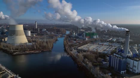 large industrial power plant emitting large amounts of smoke over the city berlin next to a river on a sunny day. dramatic aerial view flight static tripod hovering drone