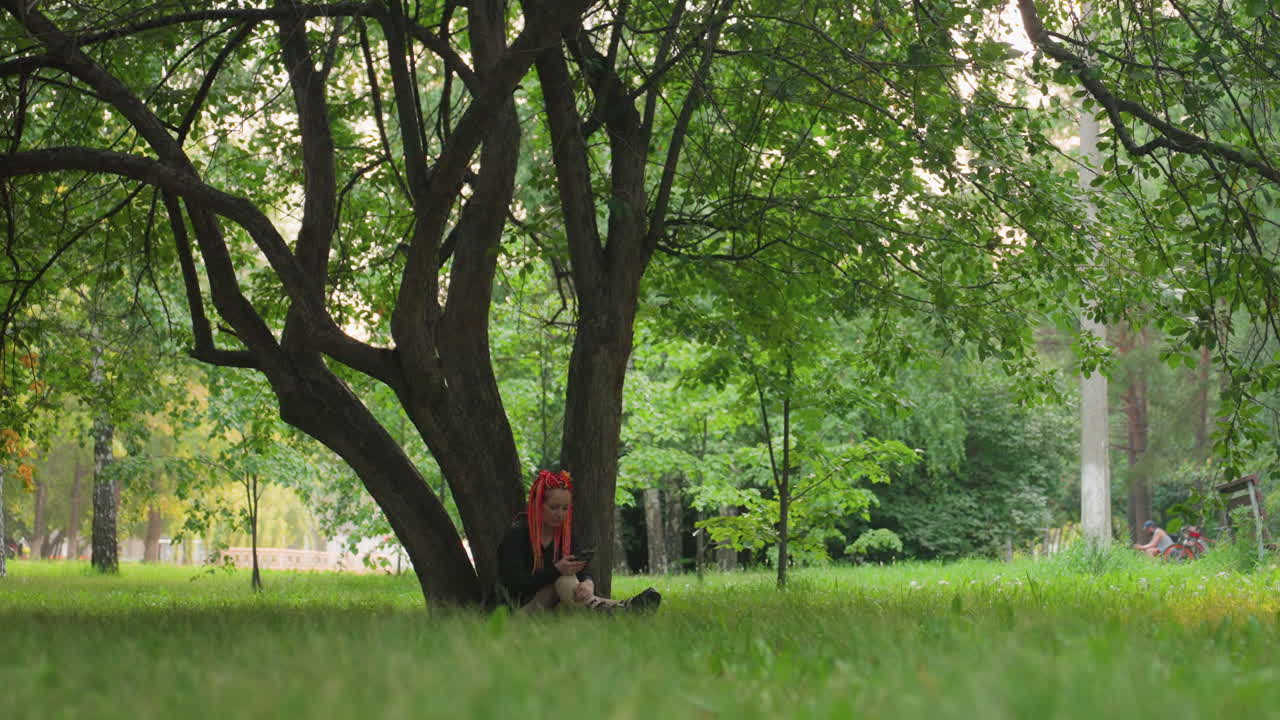 부드러운 풀밭 위에 잎이 무성한 나뭇가지 아래 누워 있는 젊은 작가, 발치에는 백팩, 나뭇잎 사이로 보이는 멀리 있는 도시 풍경, 사색에 잠긴 채 위를 올려다보는 피사체, 친밀하고 고요한 분위기