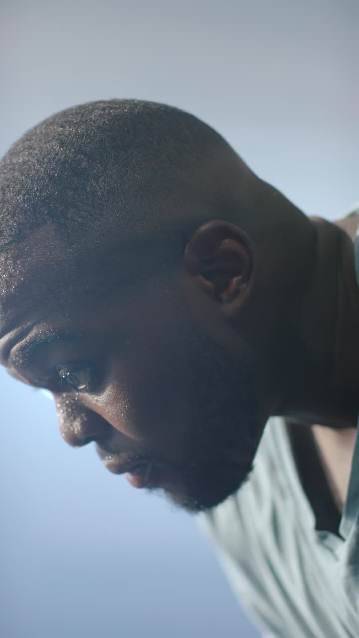 Vertical video: African man hand wiping sweat after workout breathing near backlit window light tee