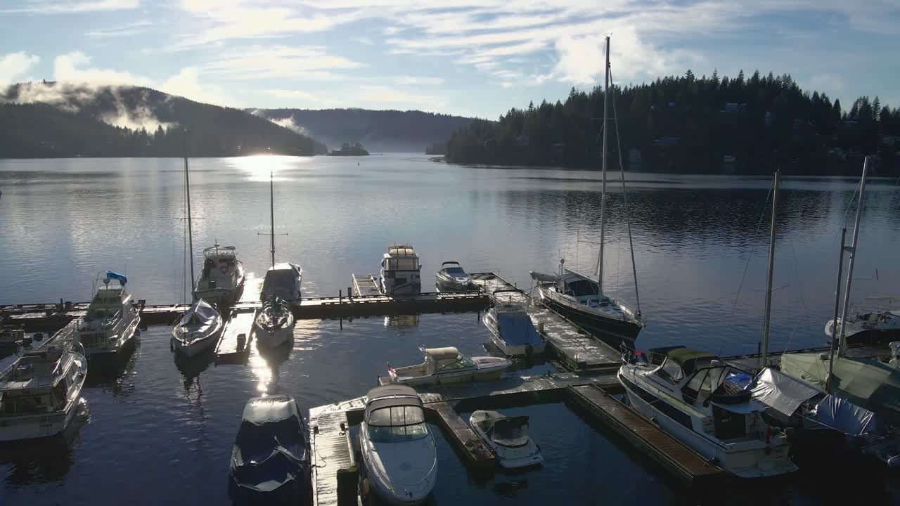 Multiple sailing yachts moored at a wooden scaffolding in a marina with islands in the Indian Arm bay at background on a sunny day