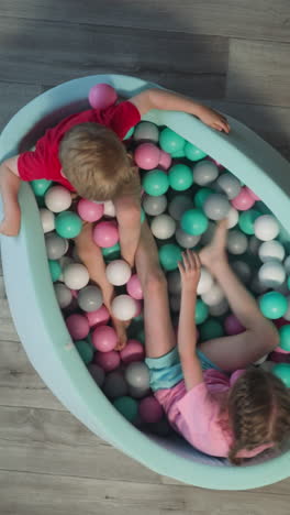 little brother and sister sit in dry pool with colorful balls. blonde girl throws plastic ball on younger boy and pink toy rolls on floor