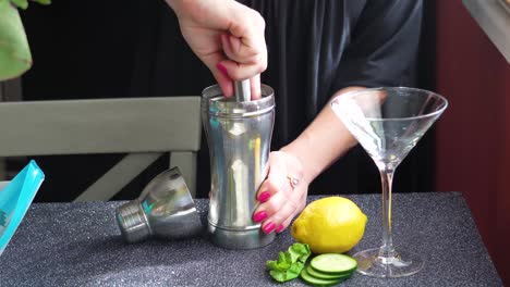 Bartender-Muddles-Cocktail-Ingredients-in-a-Silver-Cobbler-Shaker-for-a-Mixed-Drink-in-a-Martini-Glass-with-Lemon,-mint-leaves-and-Cucumber-Slices,-Closeup-of-Woman’s-Hands-on-Granite-Table