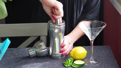 el camarero mezcla los ingredientes del cóctel en una coctelera de plata para una bebida mixta en un vaso de martini con limón, hojas de menta y rodajas de pepino, el cierre de las manos de una mujer sobre una mesa de granito