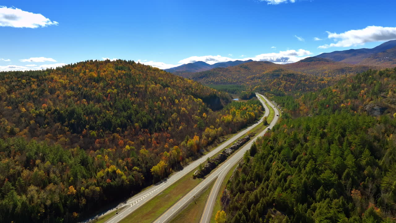 Lorries ride by the highways in the mountainous area on sunny day. Drone footage above the rocks covered with thick woods colored in green, red and yellow colors.