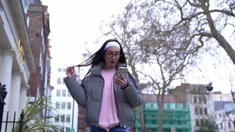 Cute-girl-walking-down-street-looking-at-phone,-holding-her-hair