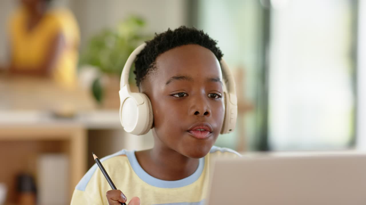 African American male child wearing headphones pointing with pencil sharing ideas to laptop at desk