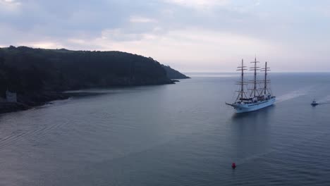 A-large-sail-powered-cruise-ship-is-arriving-in-port,-with-stunning-sun-rise-colours,-rolling-coastal-hills-and-horizon-in-the-background,-arriving-in-Dartmouth,-Devon,-United-Kingdom
