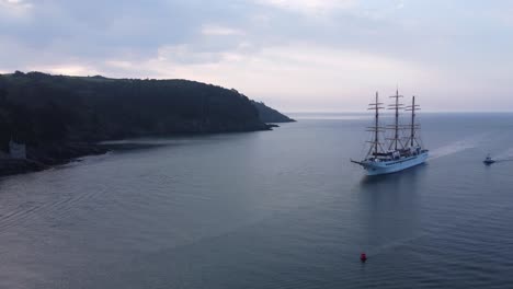 a large sail powered cruise ship is arriving in port, with stunning sun rise colours, rolling coastal hills and horizon in the background, arriving in dartmouth, devon, united kingdom