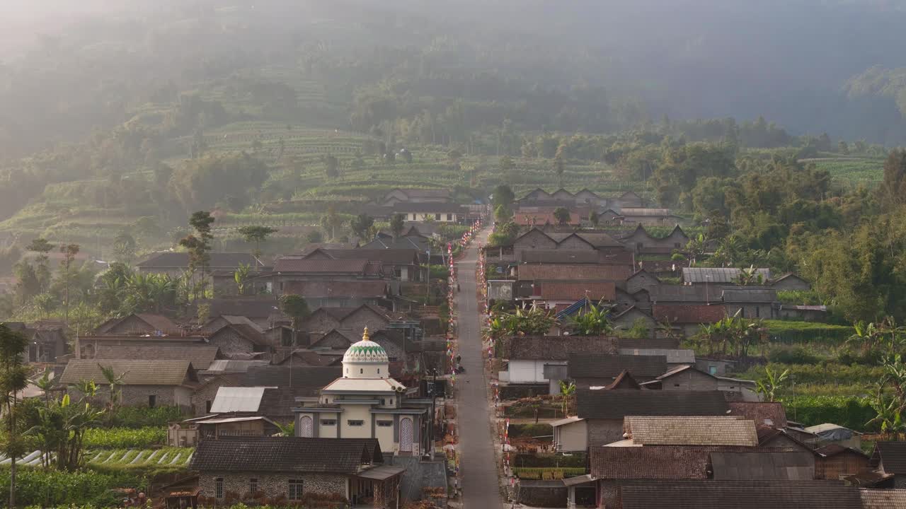 Aerial view of rural village with traditional houses and lush green farmland on a hillside under a soft morning haze. Village on the slope of Merapi volcanic mountain, Indonesia