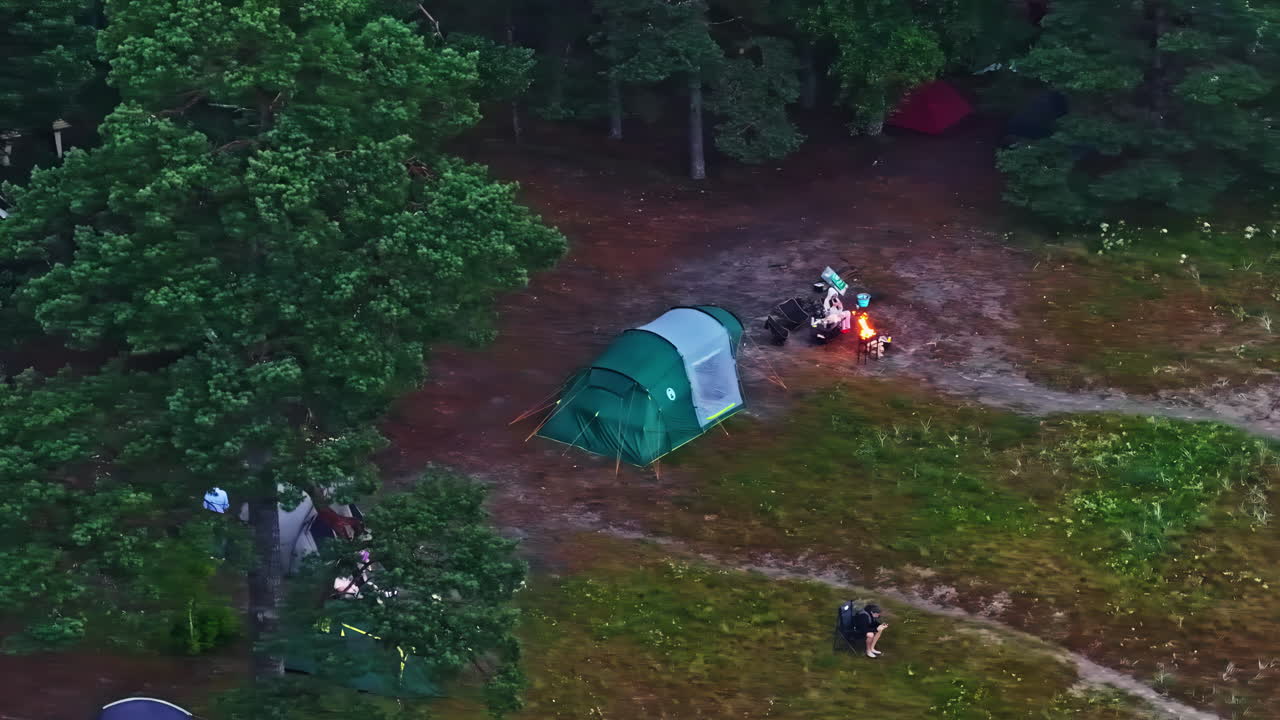Drone orbits campsite with fire pit at dusk while people walk under dim sky and smoke
