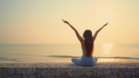 mujer joven sentada en un muro de contención cerca de la orilla estirando ambas manos mientras disfruta de la puesta de sol