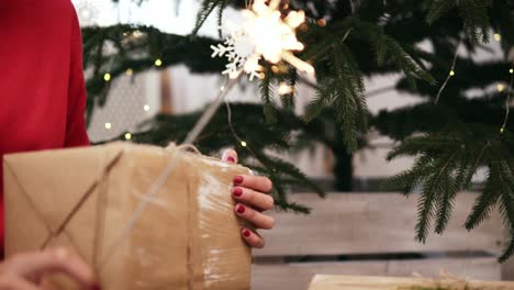 Young-smiling-woman-with-the-Christmas-tree-and-garlands-on-the-background-holding-sparklers-and-holding-a-gift-in-her-hands