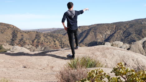 a young man on a hike in the california mountains stands at the top of a cliff overlooking the landscape in slow motion