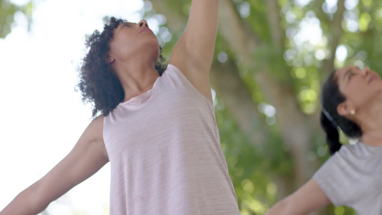 Donne diverse che seguono un'indicazione, eseguono affondi, si torcono, si allungano verso l'alto e fanno stretching su tappetini da yoga sotto gli alberi