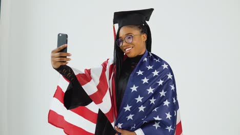 feliz estudiante afroamericana elegante en uniforme de graduado tomando una selfie sosteniendo la bandera de los estados unidos en los hombros