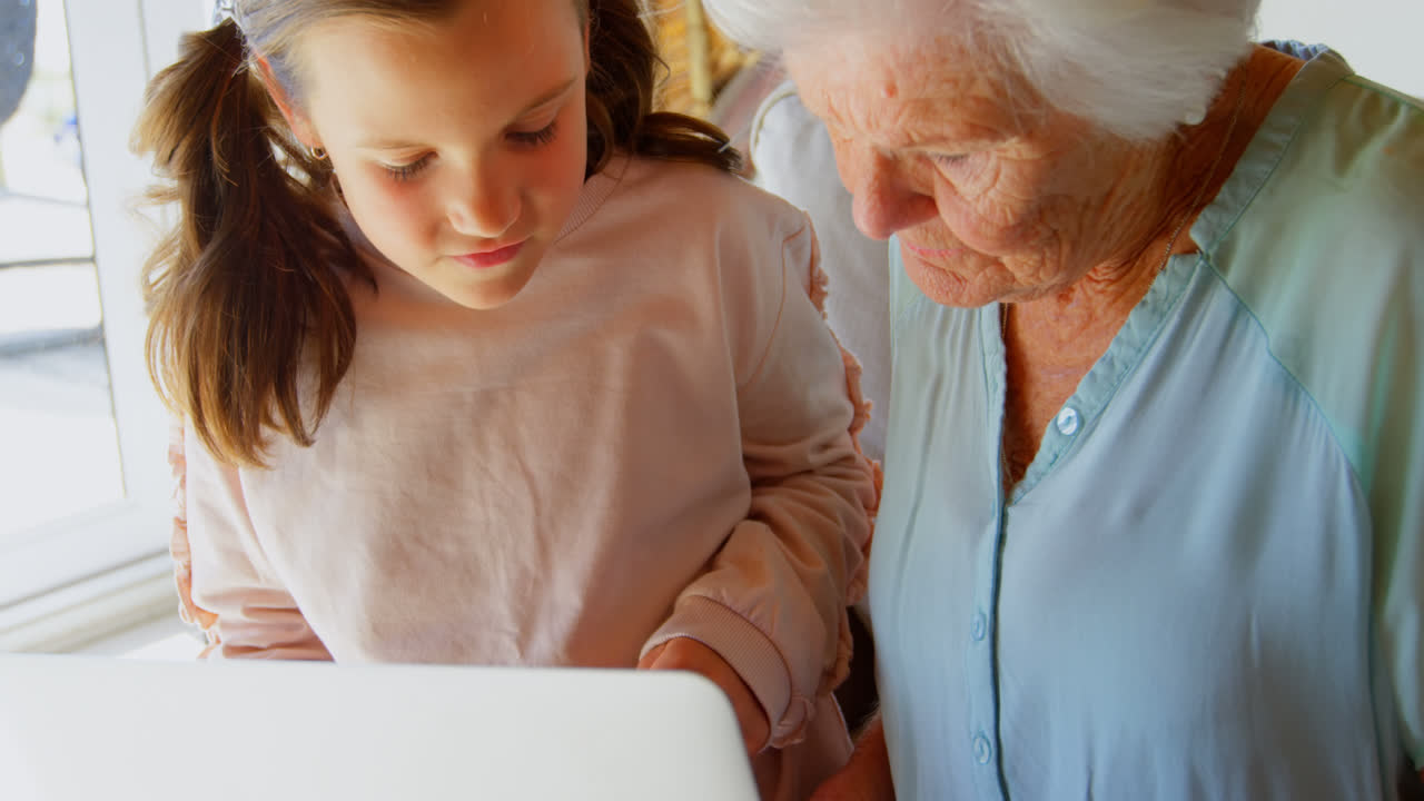 vista frontal de la abuela y la nieta caucásicas usando la computadora portátil en la sala de estar en un hogar cómodo