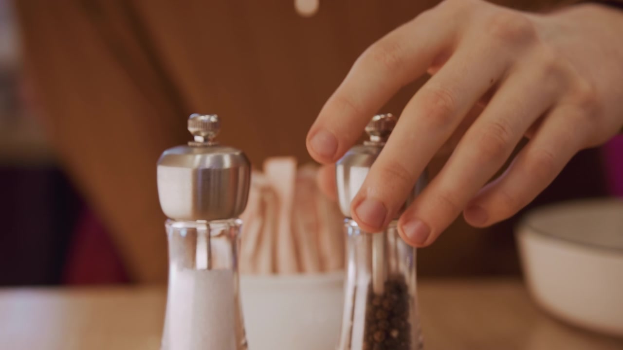 Primo piano di una mano che afferra e rimuove la pepiera davanti al sale. Lo sfondo mostra un ambiente da ristorante con bustine di zucchero sul tavolo, una ciotola, illuminazione soffusa, espressione calma e dettagli della trama.