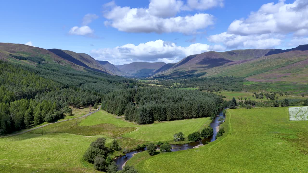 Drone glides above green valley, forest, winding river, and mountains under bright daylight skies
