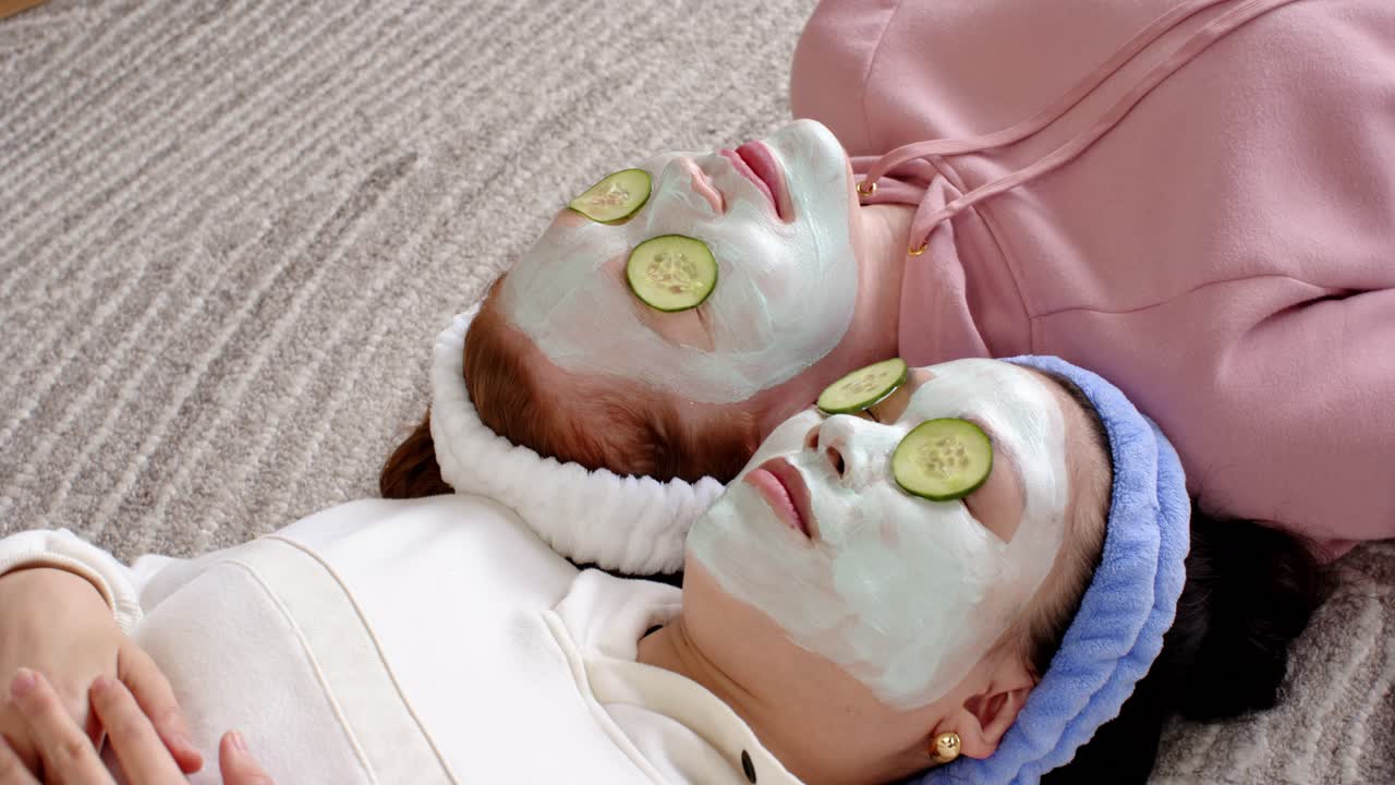 After applying masks, two women are lying on carpet near baseboard, relaxing with cucumber slices