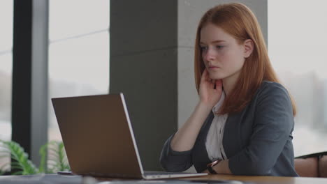 a young red-haired business woman looks thoughtfully at the screen and brainstorms. watch and think about problems looking out the window. thoughtful business woman