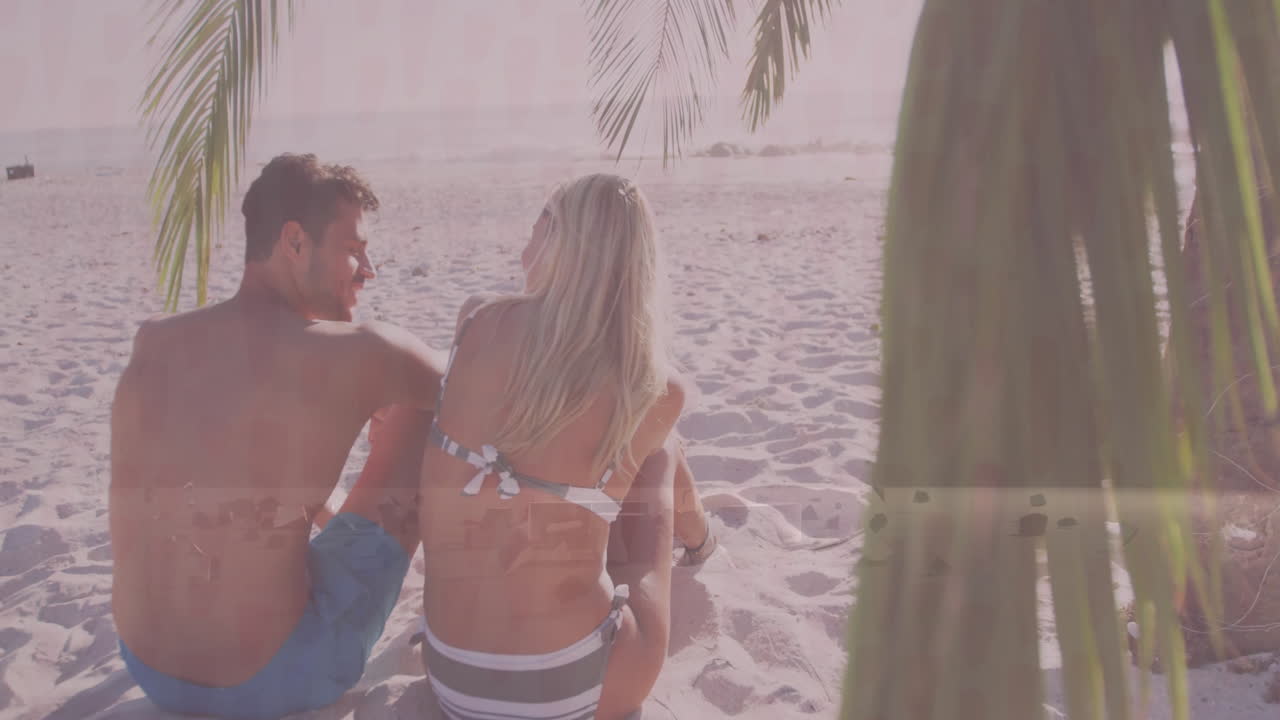 Sitting on sandy beach under palm tree, couple watching animation on tablet
