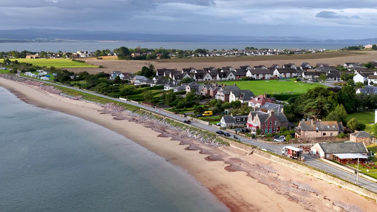 Drone glides above Rosemarkie coastline, revealing beachfront houses, sandy shore, and rural landscape