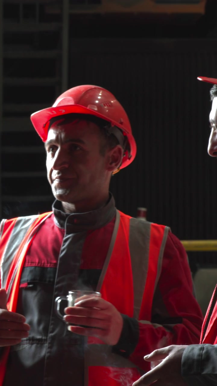 Construction Workers In Safety Vests Discussing Project Tasks Inside Dim Industrial Warehouse, Foreman