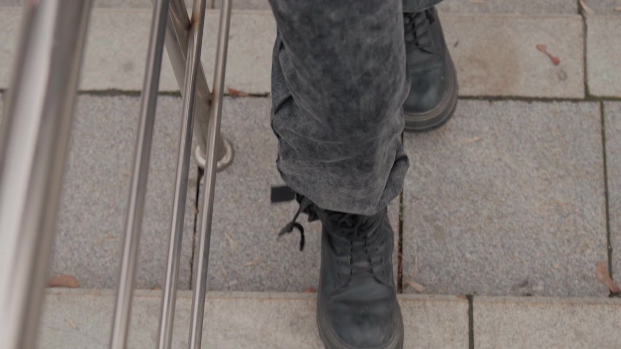 Closeup black boots walking on pavement, urban courier descending stone steps beside metal railing, worn denim pants and scuffed leather, visible laces and tread, small autumn leaves and muted tones