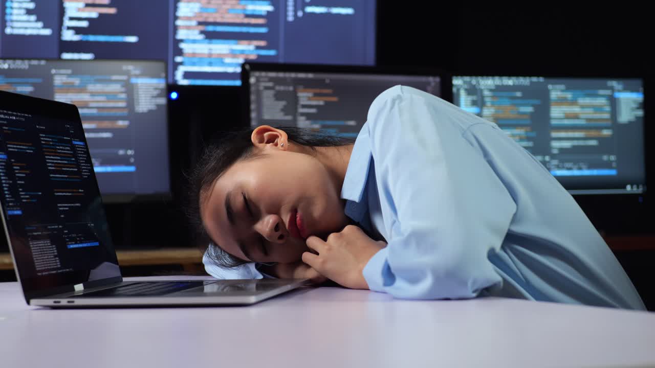 Close Up Side View Of Asian Female Programmer Sleeping While Writing Code By A Laptop Using Multiple Monitors Showing Database On Terminal Window Desktops In The Office