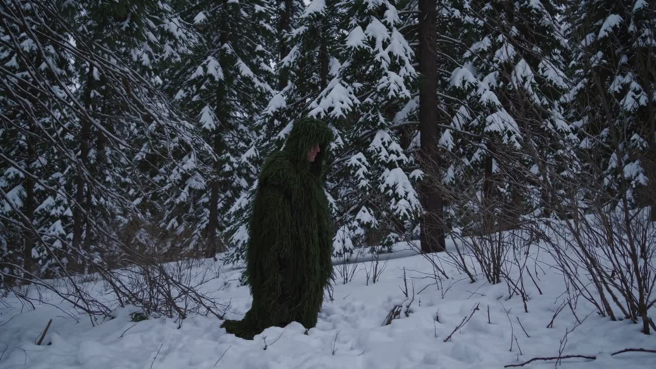 A person in a ghillie suit blends into a snowy forest. The low-angle shot captures the serene