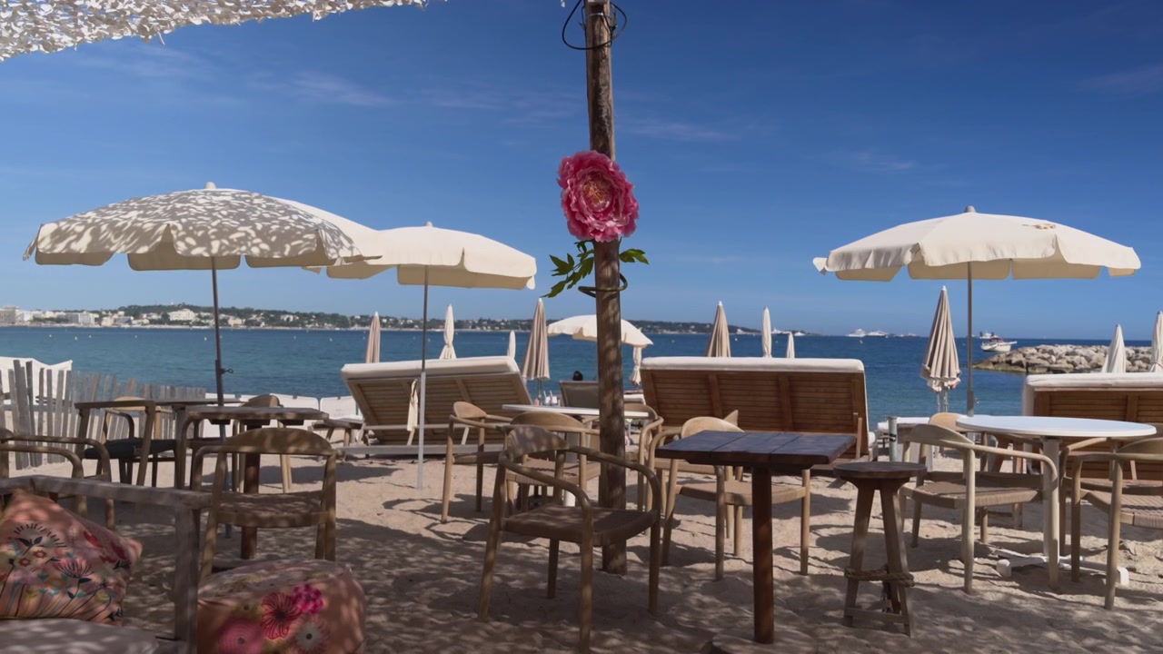 Weitläufige Aufnahmen eines Beachclubs mit Holzmöbeln, Zierblumen, Sonnenschirmen und Meerblick