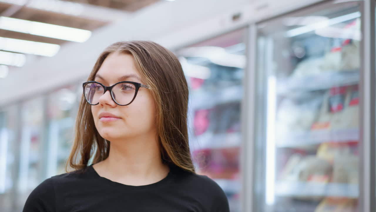 estudiante con camiseta negra y gafas camina a través de un centro comercial bien iluminado, mirando a su alrededor contemplativamente a las mercancías expuestas detrás del vidrio, la iluminación brillante mejora la atmósfera minorista moderna